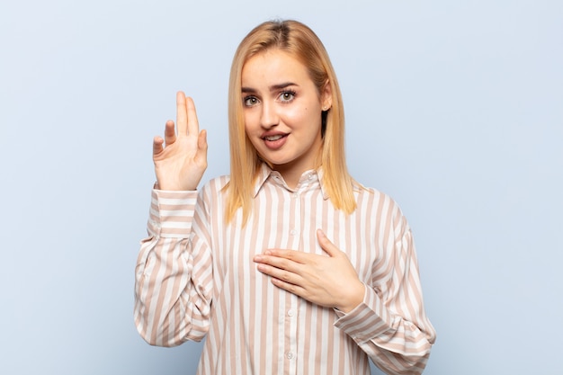 Joven mujer rubia que parece feliz, segura y digna de confianza, sonriendo y mostrando el signo de la victoria, con una actitud positiva