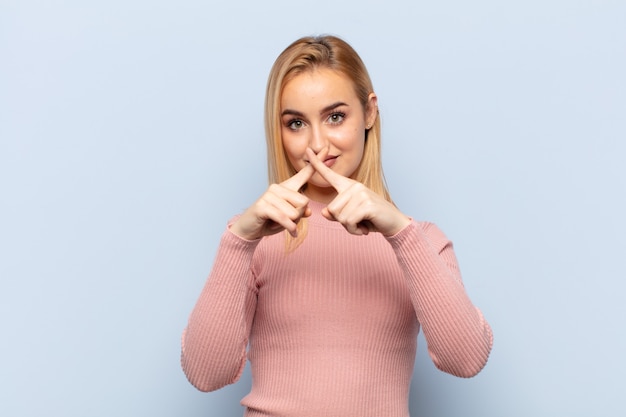 Joven mujer rubia que parece seria y disgustada con ambos dedos cruzados al frente en rechazo, pidiendo silencio