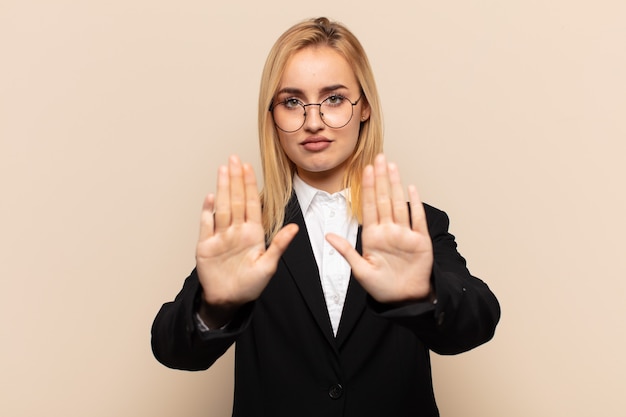 Joven mujer rubia que parece seria, infeliz, enojada y disgustada prohibiendo la entrada o diciendo que pare con ambas palmas abiertas