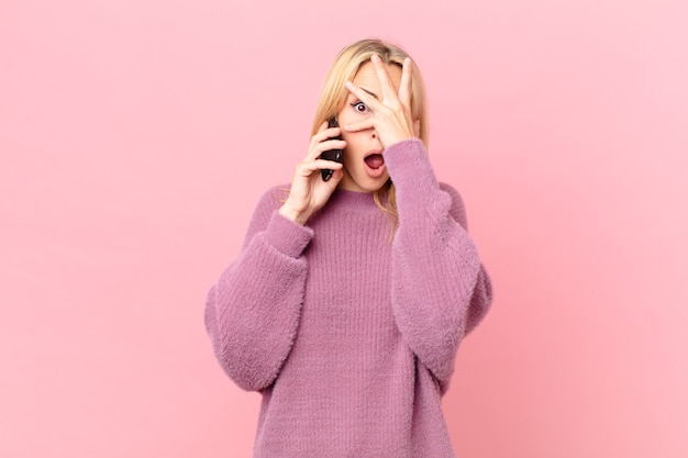 Joven mujer rubia que parece sorprendida, asustada o aterrorizada, cubriéndose la cara con la mano y hablando con un teléfono inteligente