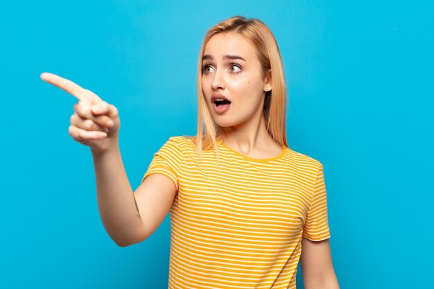 Joven mujer rubia que se siente conmocionada y sorprendida, apuntando y mirando hacia arriba con asombro con una mirada asombrada y con la boca abierta.