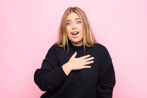 Joven mujer rubia que se siente conmocionada y sorprendida, sonriendo, tomando la mano al corazón, feliz de ser la indicada o mostrando gratitud contra la pared plana