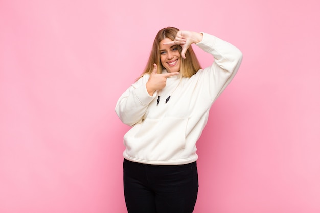 Joven mujer rubia que se siente feliz, amigable y positiva, sonriendo y haciendo un retrato o marco de fotos con las manos contra la pared plana