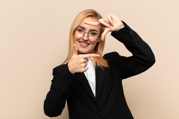 Joven mujer rubia que se siente feliz, amigable y positiva, sonriendo y haciendo un retrato o marco de fotos con las manos