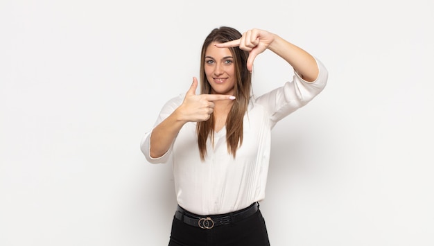 Joven mujer rubia que se siente feliz, amigable y positiva, sonriendo y haciendo un retrato o marco de fotos con las manos