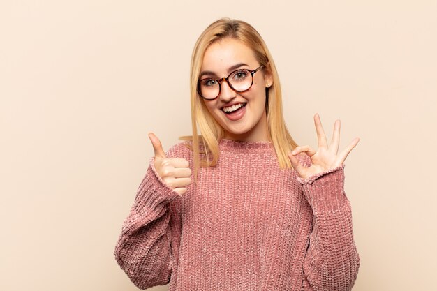 Joven mujer rubia que se siente feliz, asombrada, satisfecha y sorprendida, mostrando gestos de aprobación y pulgar hacia arriba, sonriendo