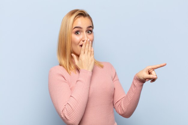 Joven mujer rubia que se siente feliz, conmocionada y sorprendida, cubriendo la boca con la mano y apuntando al espacio de copia lateral