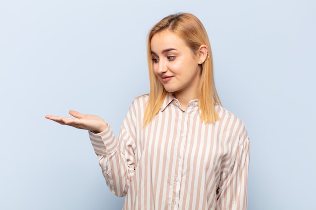 Joven mujer rubia que se siente feliz y sonriendo casualmente, mirando a un objeto o concepto sostenido en la mano al costado