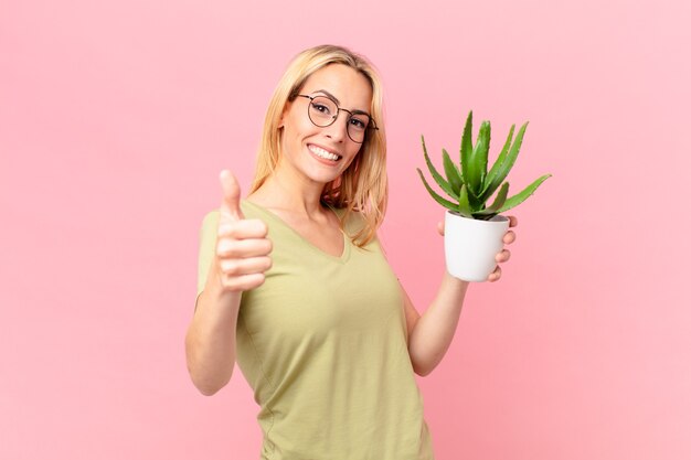 Joven mujer rubia que se siente orgullosa, sonriendo positivamente con los pulgares hacia arriba y sosteniendo un cactus