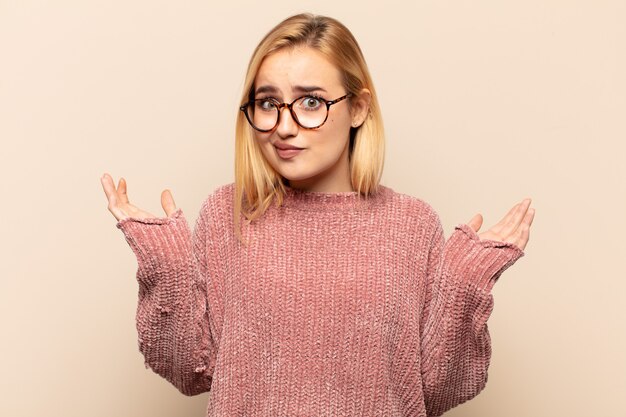 Joven mujer rubia que se siente perpleja y confundida, dudando, ponderando o eligiendo diferentes opciones con expresión divertida