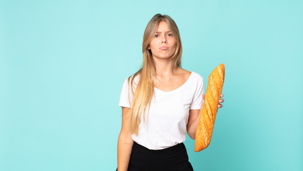 Joven mujer rubia que se siente triste y llorona con una mirada infeliz y llorando y sosteniendo una baguette de pan