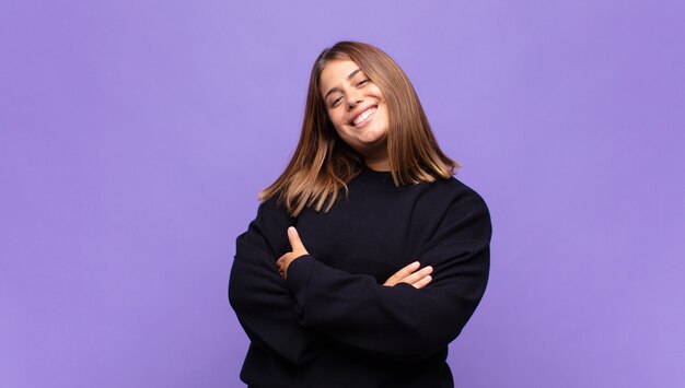 Joven mujer rubia riendo alegremente con los brazos cruzados, con una pose relajada, positiva y satisfecha