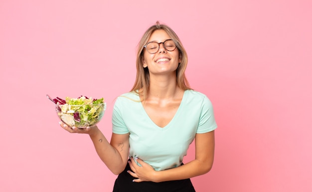 Joven mujer rubia riendo a carcajadas de una broma hilarante y sosteniendo una ensalada