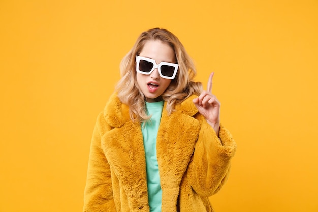 Joven mujer rubia segura de sí misma con abrigo de piel amarillo, gafas de sol oscuras posando aisladas en un retrato de estudio de fondo de pared naranja. Concepto de estilo de vida de las personas. Simulacros de espacio de copia. Señalando con el dedo índice hacia arriba.