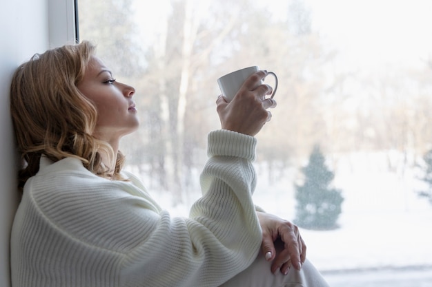 Joven mujer rubia sentada junto a la ventana con paisaje invernal. Un pasatiempo acogedor.