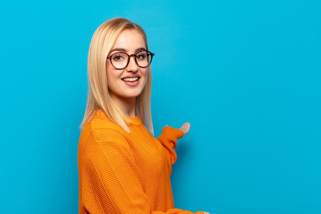 Joven mujer rubia sintiéndose feliz y alegre, sonriendo y dándote la bienvenida, invitándote con un gesto amistoso