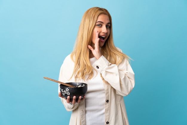 Joven mujer rubia sobre azul aislado susurrando algo mientras sostiene un plato de fideos con palillos