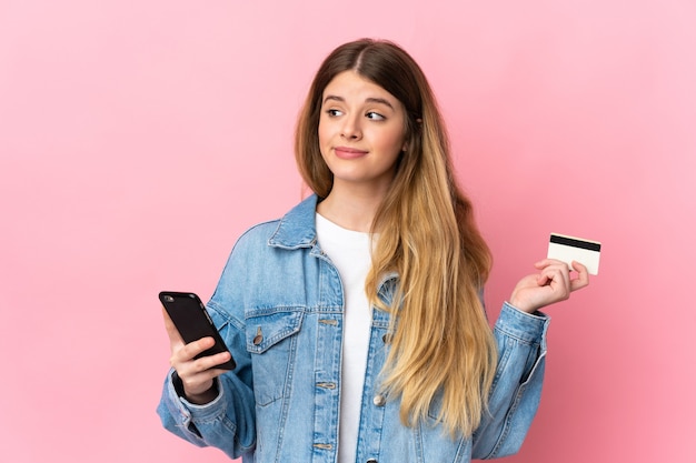 Joven mujer rubia sobre pared aislada comprando con el móvil con una tarjeta de crédito mientras piensa