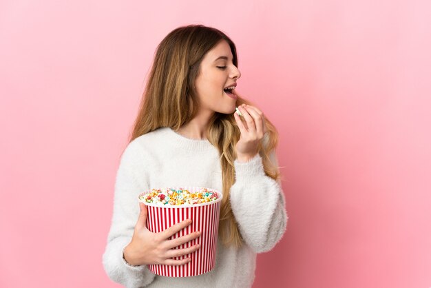 Joven mujer rubia sobre pared aislada sosteniendo un gran balde de palomitas de maíz