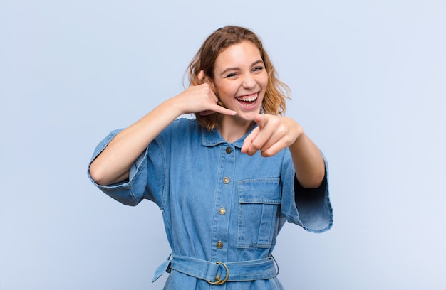 Joven mujer rubia sonriendo alegremente y apuntando a la cámara mientras hace una llamada más tarde gesto