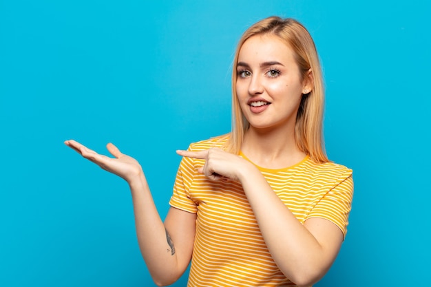 Joven mujer rubia sonriendo alegremente y señalando para copiar el espacio en la palma del costado, mostrando o publicitando un objeto