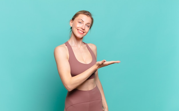 Joven mujer rubia sonriendo alegremente, sintiéndose feliz y mostrando un concepto en el espacio de la copia con la palma de la mano. concepto de deporte