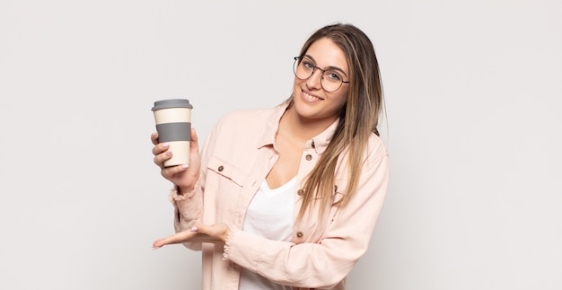 Joven mujer rubia sonriendo alegremente, sintiéndose feliz y mostrando un concepto en el espacio de la copia con la palma de la mano