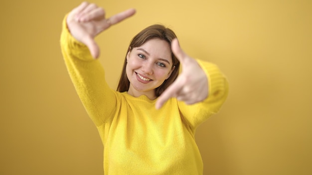 Joven mujer rubia sonriendo confiada haciendo un gesto fotográfico con las manos sobre un fondo amarillo aislado