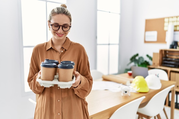 Joven mujer rubia sonriendo confiada sosteniendo café para llevar en el estudio del arquitecto