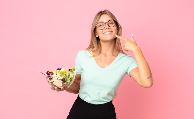 Joven mujer rubia sonriendo con confianza apuntando a su propia amplia sonrisa y sosteniendo una ensalada