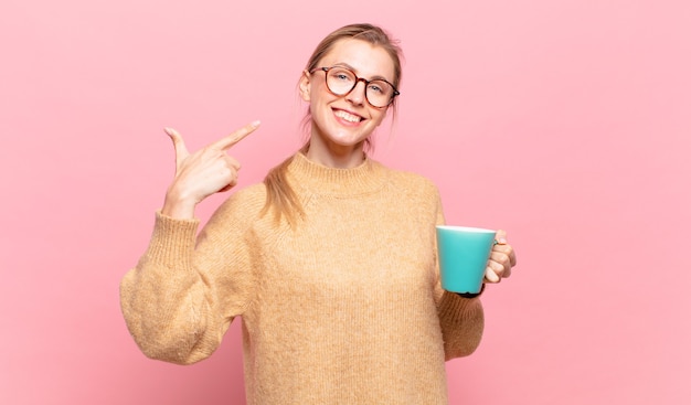 Joven mujer rubia sonriendo con confianza apuntando a su propia sonrisa amplia, actitud positiva, relajada y satisfecha. concepto de cafe