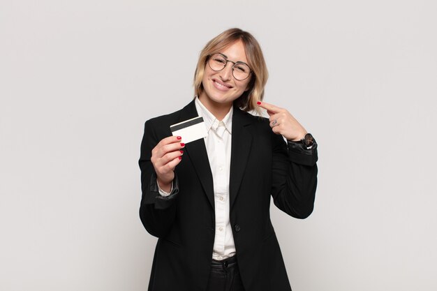 Joven mujer rubia sonriendo con confianza apuntando a su propia sonrisa amplia, actitud positiva, relajada y satisfecha