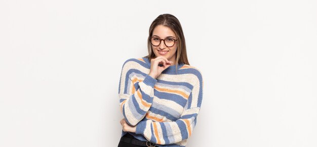 Joven mujer rubia sonriendo, disfrutando de la vida, sintiéndose feliz, amigable, satisfecho y despreocupado con la mano en la barbilla