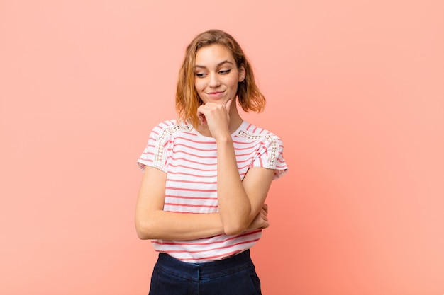 Joven mujer rubia sonriendo con una expresión feliz y segura con la mano en la barbilla, preguntándose y mirando a un lado contra la pared de color plano