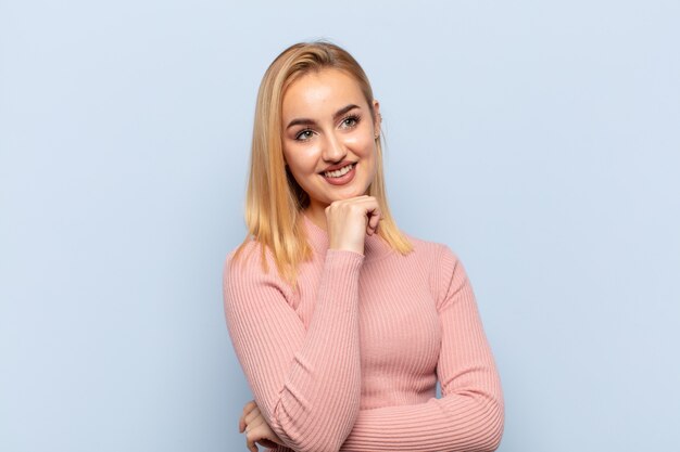 Joven mujer rubia sonriendo con una expresión feliz y segura con la mano en la barbilla, preguntándose y mirando hacia un lado