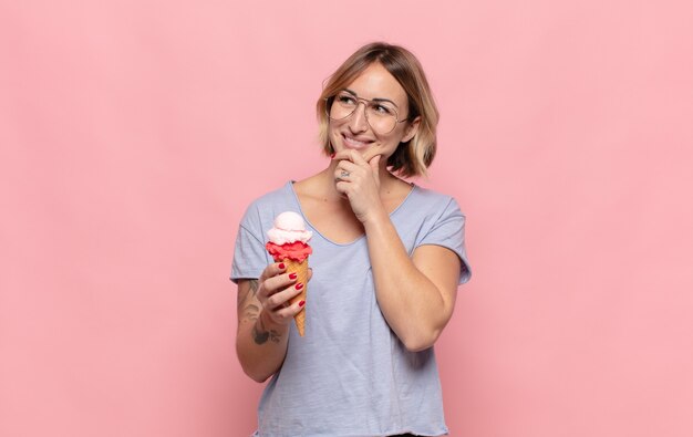 Joven mujer rubia sonriendo con una expresión feliz y segura con la mano en la barbilla, preguntándose y mirando hacia un lado