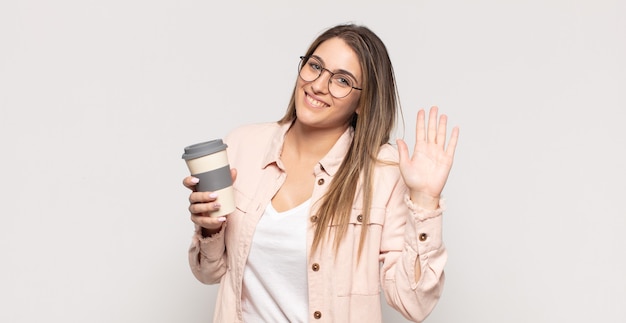 Joven mujer rubia sonriendo feliz y alegremente, saludando con la mano, dándote la bienvenida y saludándote, o diciéndote adiós