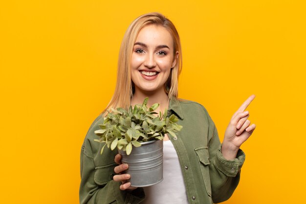 Joven mujer rubia sonriendo felizmente y apuntando hacia un lado y hacia arriba con ambas manos mostrando el objeto en el espacio de la copia