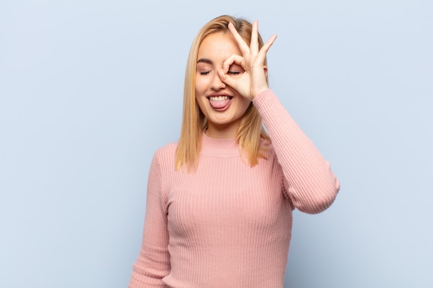 Joven mujer rubia sonriendo felizmente con cara divertida, bromeando y mirando por la mirilla, espiando secretos