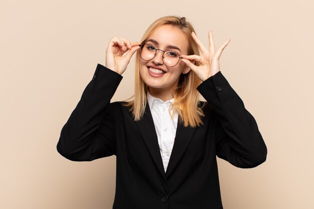 Joven mujer rubia sonriendo felizmente con cara divertida, bromeando y mirando a través de la mirilla, espiando secretos