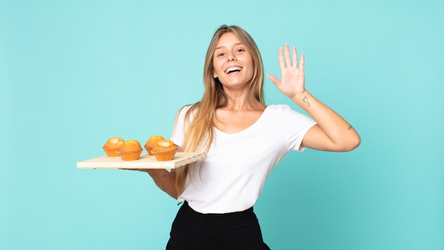 Joven mujer rubia sonriendo felizmente, saludando con la mano, dándote la bienvenida y saludándote y sosteniendo un muffins troy