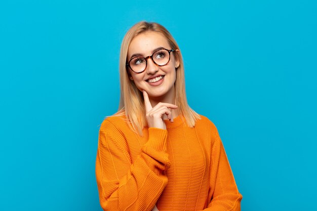 Joven mujer rubia sonriendo felizmente y soñando despierto o dudando, mirando hacia el lado