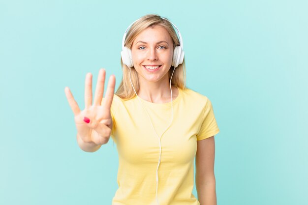 Joven mujer rubia sonriendo y mirando amigable, mostrando el número cuatro y escuchando música.