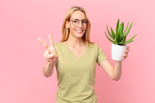 Joven mujer rubia sonriendo y mirando amigable, mostrando el número dos y sosteniendo un cactus