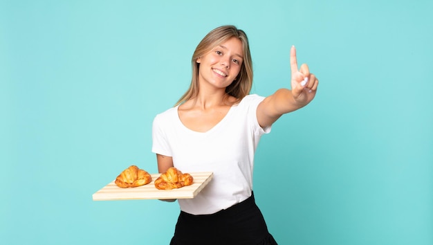 Joven mujer rubia sonriendo y mirando amigable, mostrando el número uno y sosteniendo una bandeja de croissant