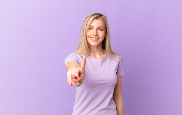 Joven mujer rubia sonriendo y mirando amigable, mostrando el número uno