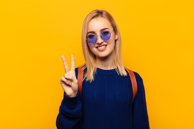 Joven mujer rubia sonriendo y mirando amistosamente, mostrando el número dos o el segundo con la mano hacia adelante, contando hacia atrás