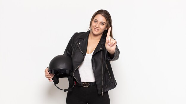 Joven mujer rubia sonriendo y mirando amistosamente, mostrando el número uno o el primero con la mano hacia adelante, contando hacia atrás