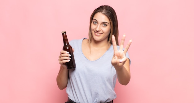 Joven mujer rubia sonriendo y mirando amistosamente, mostrando el número tres o tercero con la mano hacia adelante