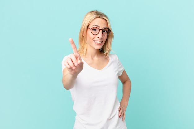 Joven mujer rubia sonriendo con orgullo y confianza haciendo el número uno